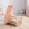 A wooden rocking doll cradle with a floral canopy and bedding, set in a child's room with soft furnishings. Produktbild-Vorschau