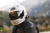 A person wearing a white motorcycle helmet and black leather jacket, with a blurred mountainous background. Anteprima dell’immagine prodotto