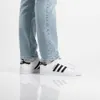 A person wearing Adidas Superstar II shoes and light blue jeans, standing on a white reflective surface. Voir les images du produit