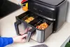 A person's hand is pulling out the drawers of a dual-compartment air fryer, which contains food in glass containers. Náhľad obrázku produktu