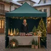 A woman stands behind a market stall decorated for Christmas, selling jars of preserves. Anteprima dell’immagine prodotto