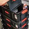 A person's hand wearing a glove is opening the lid of a stack of black Qbrick System transport boxes. One box contains a spray can and other items. Náhled obrázku produktu