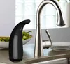 A black automatic soap dispenser and a silver faucet are on a kitchen counter next to a bowl of grapes and a stack of plates. Náhled obrázku produktu