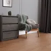 A chest of drawers with a cactus and a framed picture on top, next to a chair with a blanket and pillow, in a room with wooden flooring and dark curtains. Náhled obrázku produktu