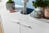A close-up view of a white bathroom cabinet with two drawers, featuring decorative handles. Several items are placed on top of the cabinet, including a small potted plant, a lipstick, a candle, and another decorative candle. Náhľad obrázku produktu