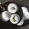 A collection of white ceramic plates and bowls arranged on a black surface, with candles and a dried flower as decorative elements. Voir les images du produit