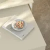 A close-up shot of a small marble-patterned bowl filled with pistachios, placed on top of a book, which is on a cream-colored surface. The background shows a textured carpet. Voir les images du produit