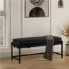 A black bench with a throw blanket draped over it, placed in a well-lit room with a window and artwork on the wall. Produktbild-Vorschau