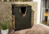 A firewood storage unit with a cover is shown on a patio next to a brick wall and a window with a fireplace inside. Produktbild-Vorschau