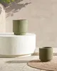 Two green planters with a ribbed texture are displayed outdoors on a patio. One planter is on a white pedestal, and the other is on a round jute rug. The background is a textured beige wall with shadows from foliage. Podgląd zdjęcia produktu