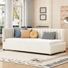 A beige daybed with several decorative pillows is shown in a well-lit living room with large windows and sheer curtains. Náhľad obrázku produktu