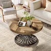 A round wooden coffee table with a metal base is placed on a rug in a living room setting, next to a sofa and armchair. Voir les images du produit