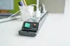 A close-up of a black power strip with a green on/off switch and an orange reset button, placed on a white desk. Produktbild-Vorschau