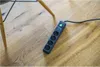 A black power strip with four outlets is lying on a wooden floor next to the legs of a chair. Produktbild-Vorschau