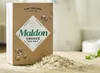 A box of Maldon Smoked Sea Salt flakes is shown on a wooden surface, with a pile of salt in front of it. Náhled obrázku produktu