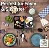 A raclette set is laid out on a wooden table with various food items, including grilled salmon, skewers, and a fried egg, ready for a meal. Produktbild-Vorschau