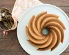 A bundt cake is presented on a light blue plate, with a copper cup filled with silverware and a pink cloth with gold polka dots in the background, all set on a wooden surface. Podgląd zdjęcia produktu