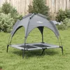 A gray dog bed with a canopy is set up on a grassy lawn in front of a wooden fence and some bushes. Náhled obrázku produktu