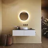 A modern bathroom vanity with a round illuminated mirror, sink, and storage drawers. A laundry basket with a towel is placed to the left, and a large plant is to the right. Podgląd zdjęcia produktu