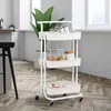 A white three-tiered rolling cart filled with kitchen items, placed on a wooden floor in a bright kitchen setting. Voir les images du produit