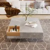 A modern living room scene featuring a square coffee table with a built-in wooden storage box, placed on a geometric rug in front of a plush white sofa. Produktbild-Vorschau
