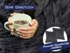 A person holding a mug of hot beverage wrapped in a dark gray plush blanket. Produktbild-Vorschau