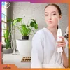 A woman in a white robe is holding a skin treatment device in a bathroom with plants and a bathtub. Produktbild-Vorschau