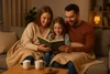 A family is reading a book together on a couch, covered by blankets. Produktbild-Vorschau