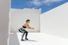 A woman is doing squats with a resistance band around her thighs in an outdoor courtyard with white walls and a blue sky. Anteprima dell’immagine prodotto
