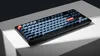 A black mechanical keyboard with blue and orange keycaps is displayed on a white surface with dramatic lighting. Náhľad obrázku produktu