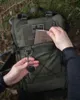 A person's hands are shown interacting with a green backpack in an outdoor, wooded setting. Produktbild-Vorschau