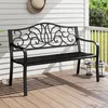 A black metal garden bench with decorative scrollwork on the backrest is placed on a wooden deck in front of a white-paneled wall. Podgląd zdjęcia produktu