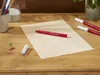 A red marker pen lies on a piece of paper on a wooden desk, with other markers and paperclips scattered around. Produktbild-Vorschau