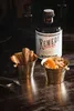 A bottle of Remedy Spiced Rum is displayed on a dark wooden surface, alongside two small metal buckets. One bucket contains cinnamon sticks, and the other holds a garnish, possibly for a drink. The scene suggests a sophisticated or rustic setting for enjoy Náhled obrázku produktu
