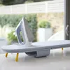 A white and blue iron rests on a gray ironing board with yellow legs, placed on a white surface. The background shows a blurred outdoor scene with greenery and a white fence. Náhľad obrázku produktu