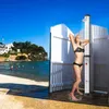 A woman is showering outdoors on a beach next to a white changing room structure. Náhľad obrázku produktu