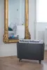 A dark gray magazine rack with magazines inside, placed on a wooden floor in front of a large ornate gold mirror. Podgląd zdjęcia produktu