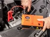 A person is using a jump starter unit to start a car battery under the hood of a car. Náhled obrázku produktu