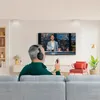 A man wearing headphones is sitting on a couch, watching TV. The TV displays a news anchor. There is a red chair and a plant in the background. Produktbild-Vorschau