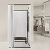 A freestanding changing room with white curtains and a dark bench inside, set against a blurred background of a retail store. Produktbild-Vorschau