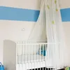A white crib with a sheer canopy and a blue stuffed toy inside, set against a wall with a blue stripe. Voir les images du produit