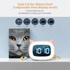 A pink cat-ear alarm clock is displayed on a shelf next to a book titled 'The Cat Book'. The clock has a digital display showing '10:10' and a soft light around the screen. Above the clock, there are circular graphics illustrating different brightness leve Produktbild-Vorschau