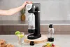 A person is using a soda maker in a kitchen setting, with a glass of sparkling water and fruit nearby. Náhled obrázku produktu
