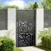A black metal garden gate with a floral cutout design, set between two white brick pillars and flanked by greenery. Anteprima dell’immagine prodotto
