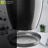 A close-up of a black plant pot and saucer, with a blurred background showing a modern kitchen and a white planter with a snake plant. Podgląd zdjęcia produktu