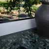 A close-up view of a window sill with a decorative vase and a view of a garden through the window. Náhled obrázku produktu