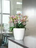 A white planter with pink and yellow orchids sits on a dark countertop, with a blurred background of a dining area and sheer curtains. Voir les images du produit