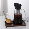 A cold brew coffee maker with a glass carafe and stainless steel filter, placed on a wooden tray with a pastry and coffee beans, next to a glass cup with a spoon. Náhled obrázku produktu