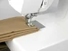 A close-up view of a sewing machine needle and presser foot positioned over folded tan fabric. Produktbild-Vorschau