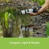 A person is holding a camera lens over a body of water with a plant growing in it. The reflection of the plant and lens is visible in the water. Produktbild-Vorschau